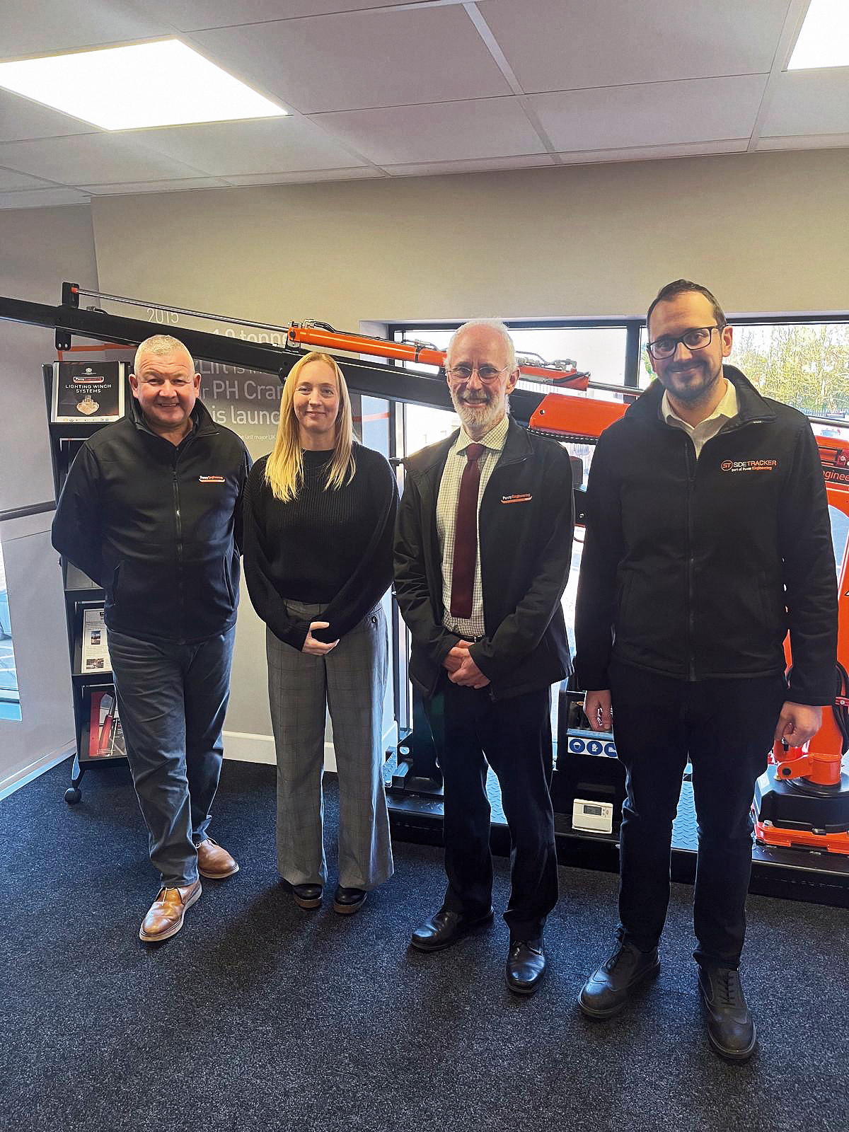 Four people stand indoors in front of orange construction equipment, dressed in business or business-casual attire, smiling at the camera. The setting appears to be an office or showroom.