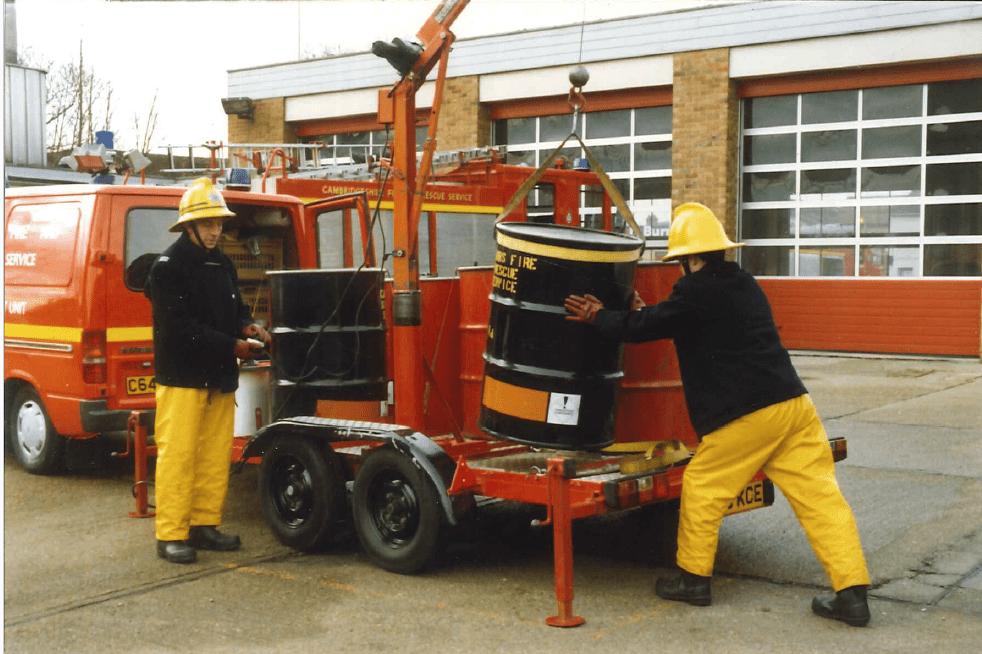 Two workers in yellow rain gear and helmets use a small crane to lift a barrel onto a trailer beside a red emergency response van, in front of a building with large garage doors.