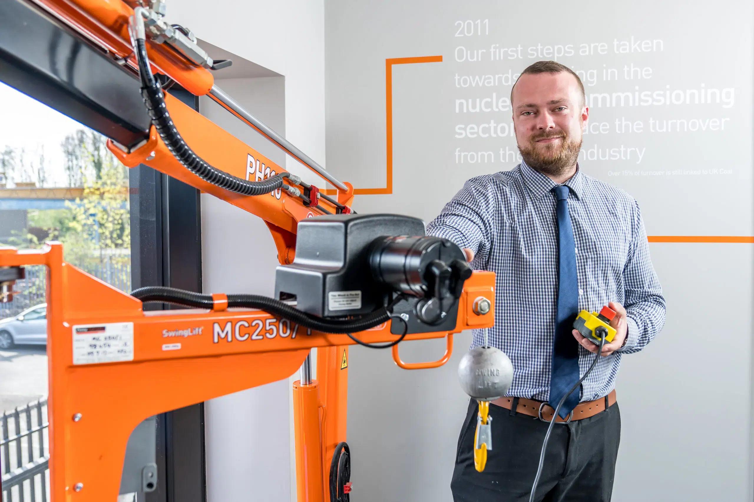 A man in a checked shirt and tie operates an orange industrial lifting machine with a control panel, standing indoors next to a wall with writing and a window showing trees outside.