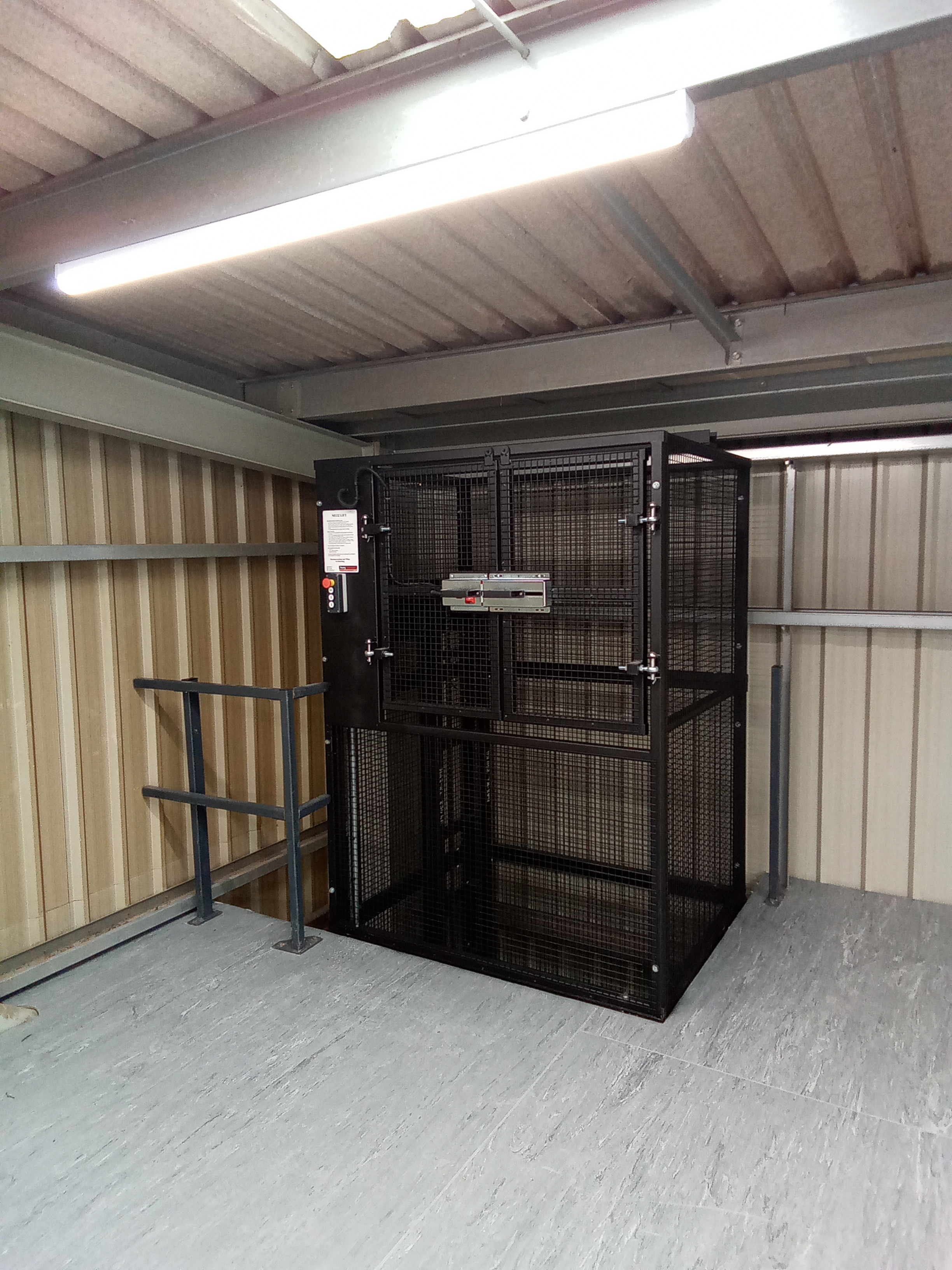 A black, metal mesh goods lift is installed in the corner of a warehouse mezzanine, surrounded by metal railings and corrugated walls, under fluorescent lighting.