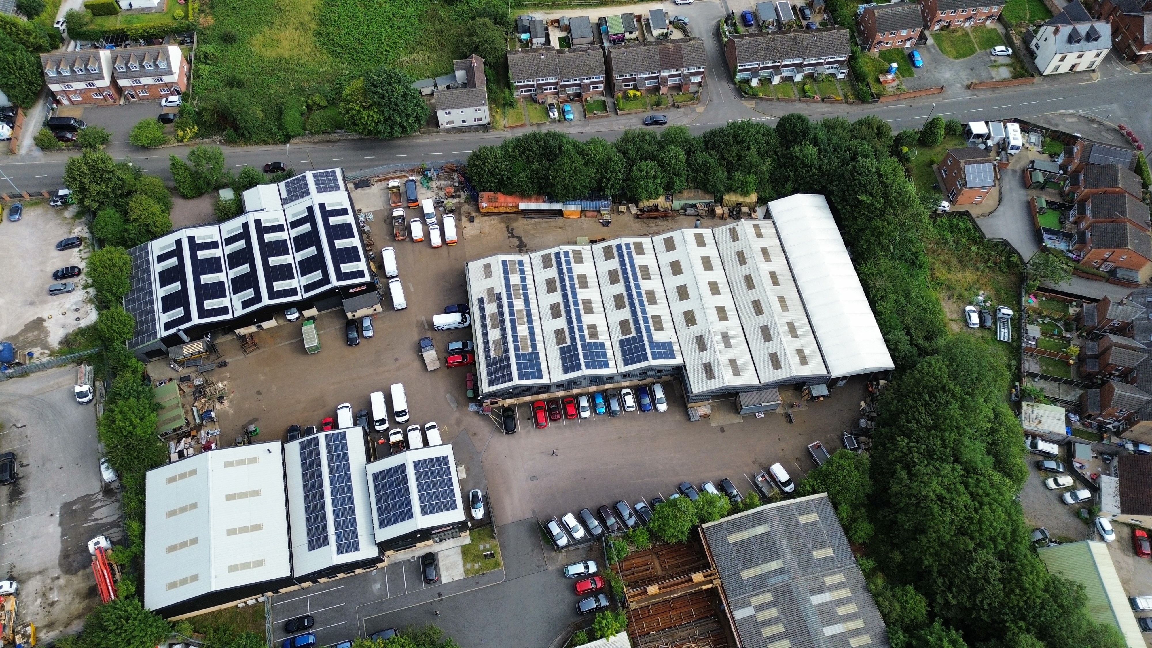 Aerial view of several large industrial buildings with solar panels on their roofs, surrounded by parked cars and trees, near a residential neighbourhood with houses and roads.