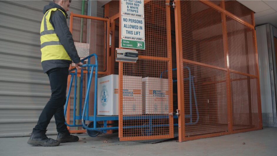 A person in a yellow safety vest pushes a blue trolley with white containers into a gated industrial lift. The lift is enclosed with orange metal fencing, and a sign restricts access to authorised personnel only.