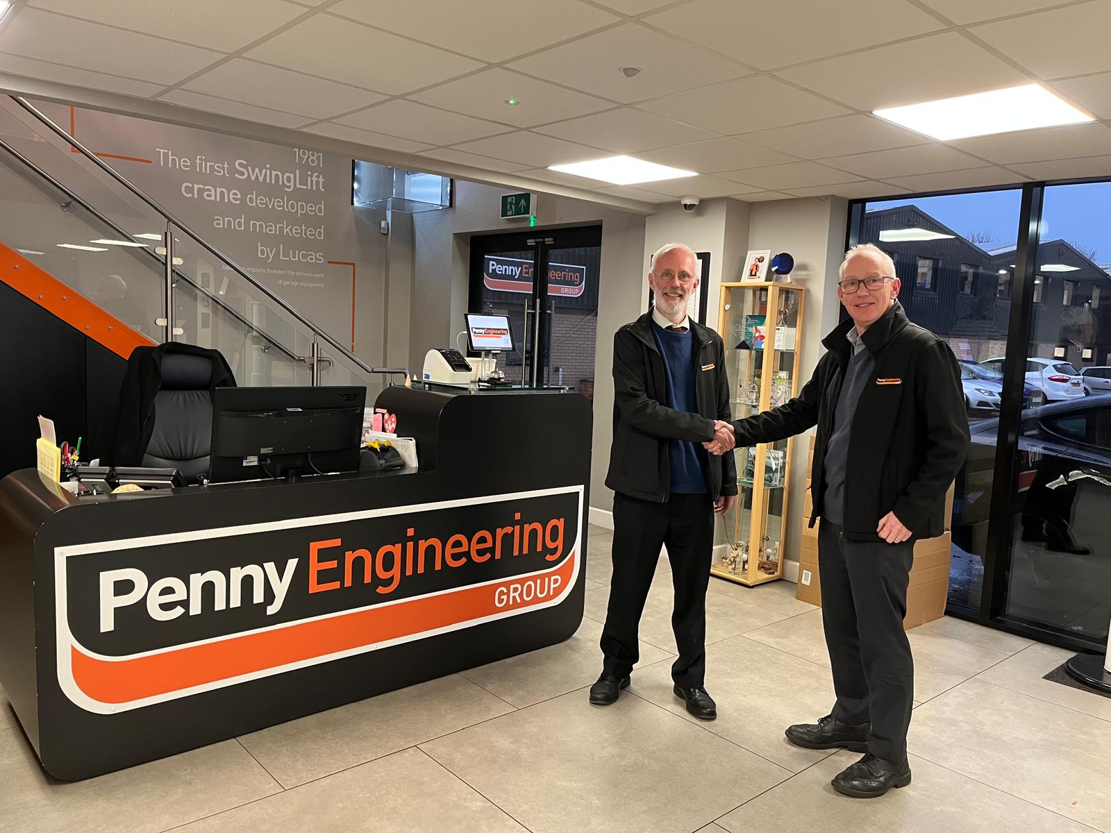 Two people stand behind a reception desk at an indoor office or shop. The desk has a sign with text, and shelves with various items are visible in the background. The image appears distorted.