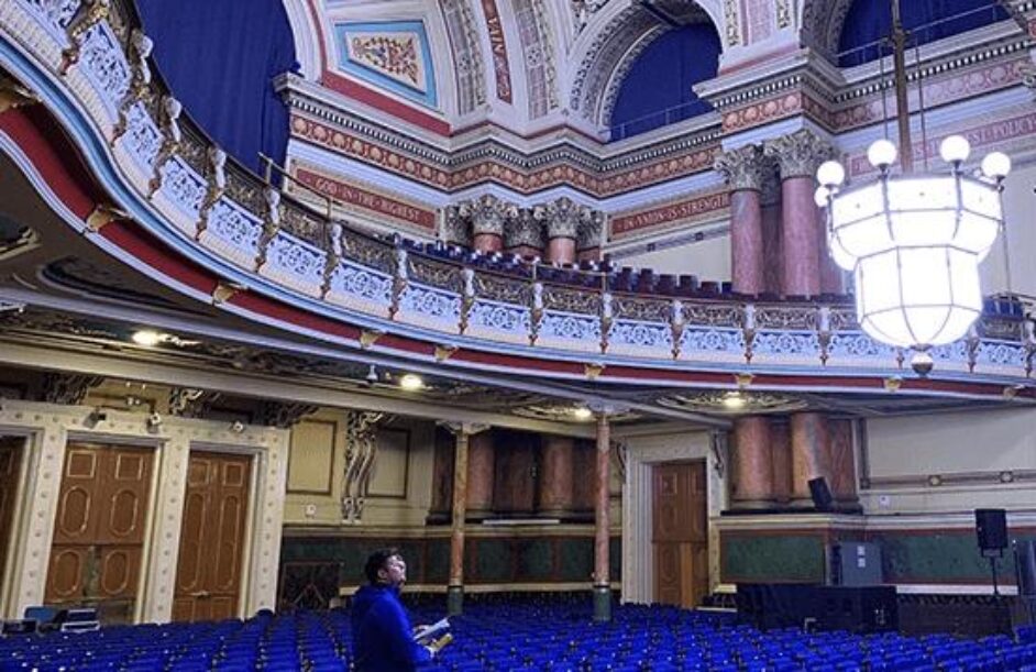 A person stands in an ornate, empty auditorium with blue seats, decorative columns, and intricate moulding. A large chandelier hangs from the high ceiling, and a balcony with detailed railings lines the upper wall.